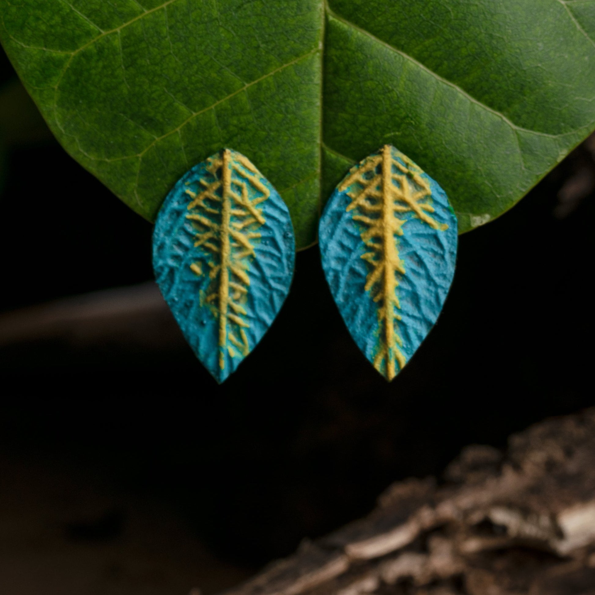 Teal leaf-shaped earrings with golden vein accents are suspended gracefully against a green leaf backdrop, creating an eye-catching contrast.