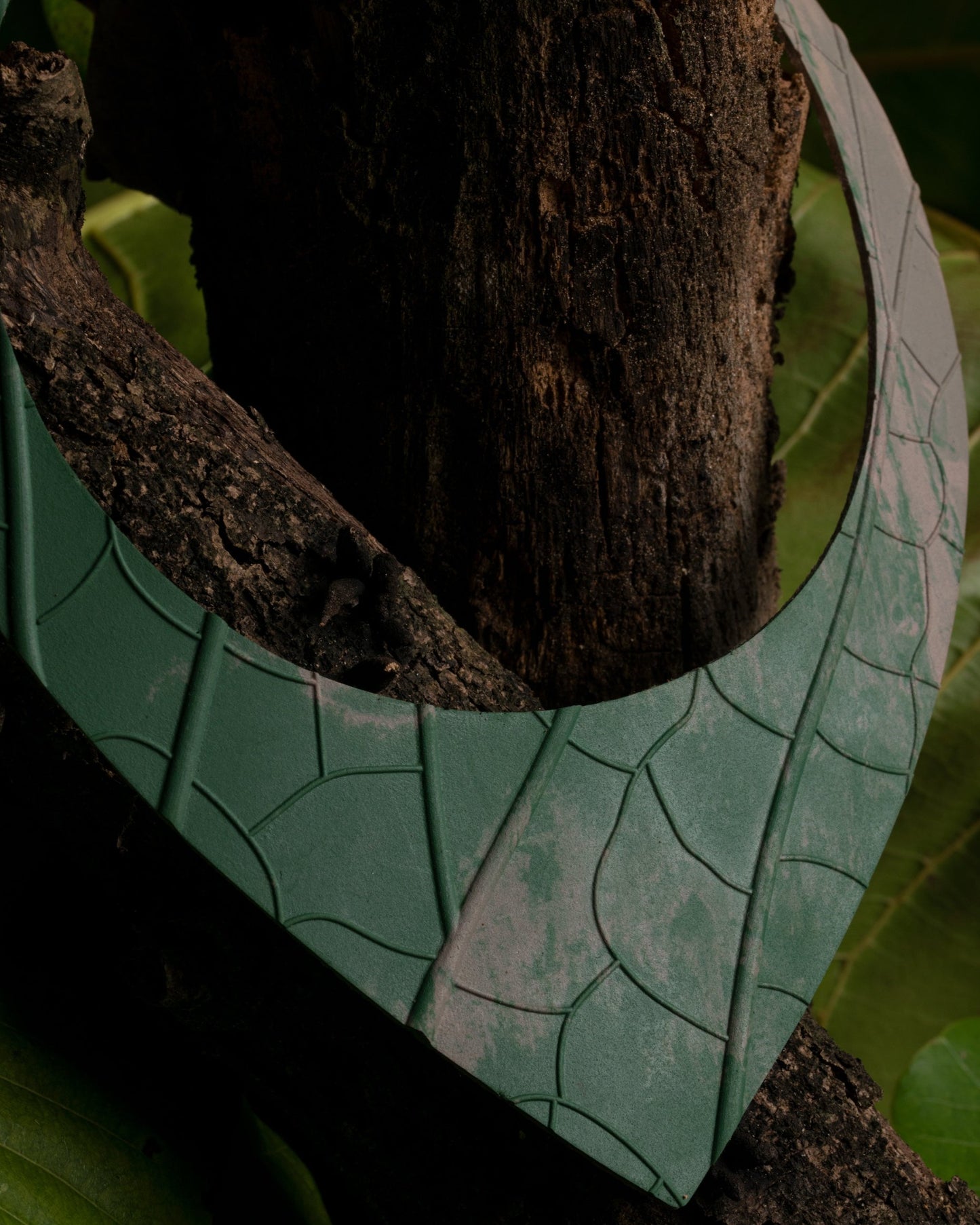 Artisanal 'Shield' necklace rests against a tree, crafted from textured Amazonian rubber in shades of matte green, highlighted by subtle açaí seed fiber patterns.