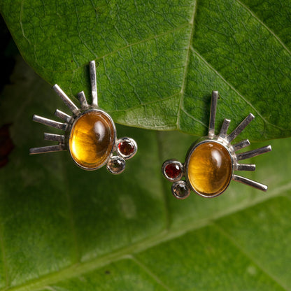 Silver earrings with tucupi gemstones, radiating a sunbeam design, nestled against a vibrant green leaf backdrop.