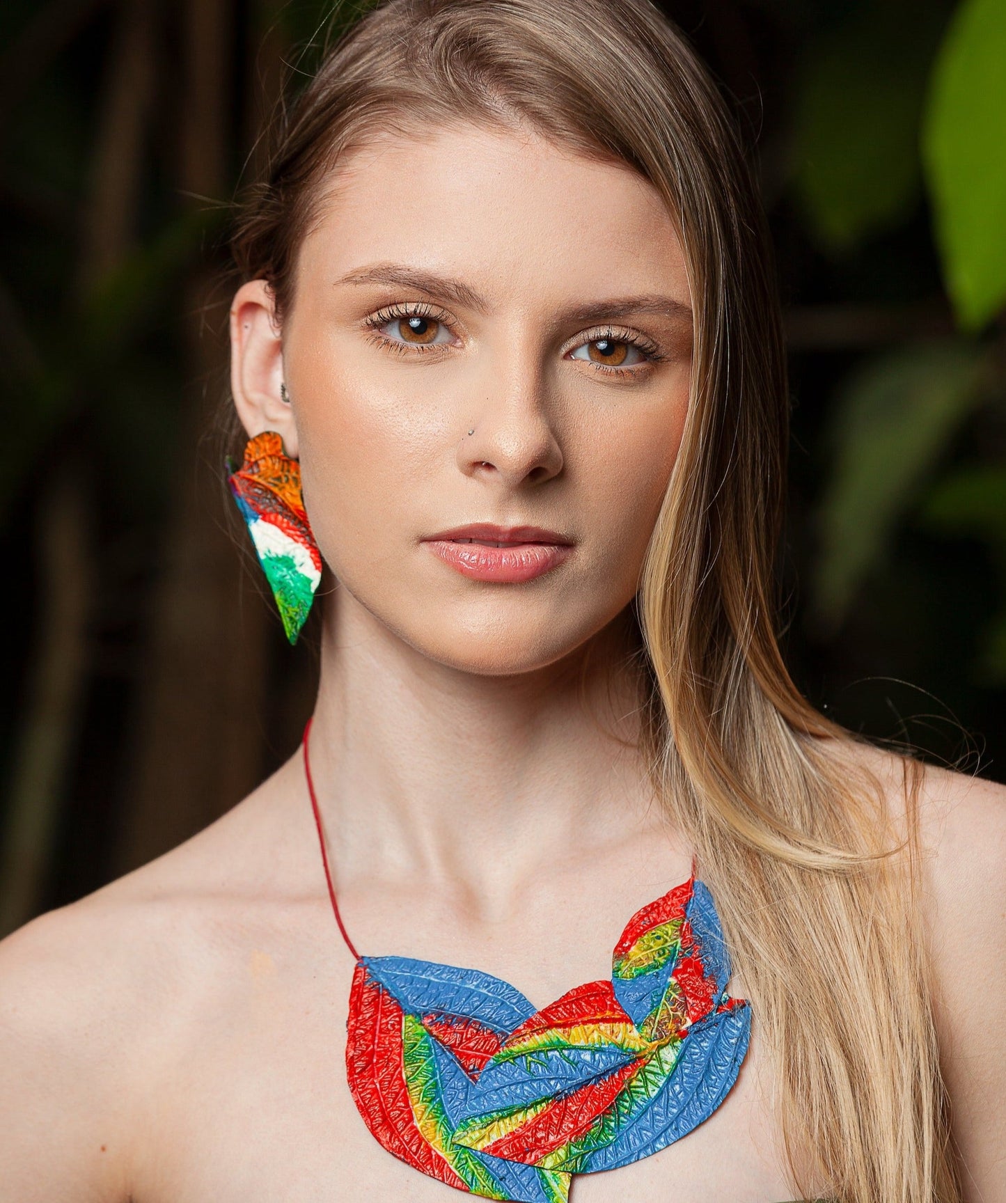 Woman models vibrant leaf necklace & earring. Handcrafted from natural rubber & açaí fibers, they feature red, green, blue leaves with detailed veins. Natural setting hints at inspiration.